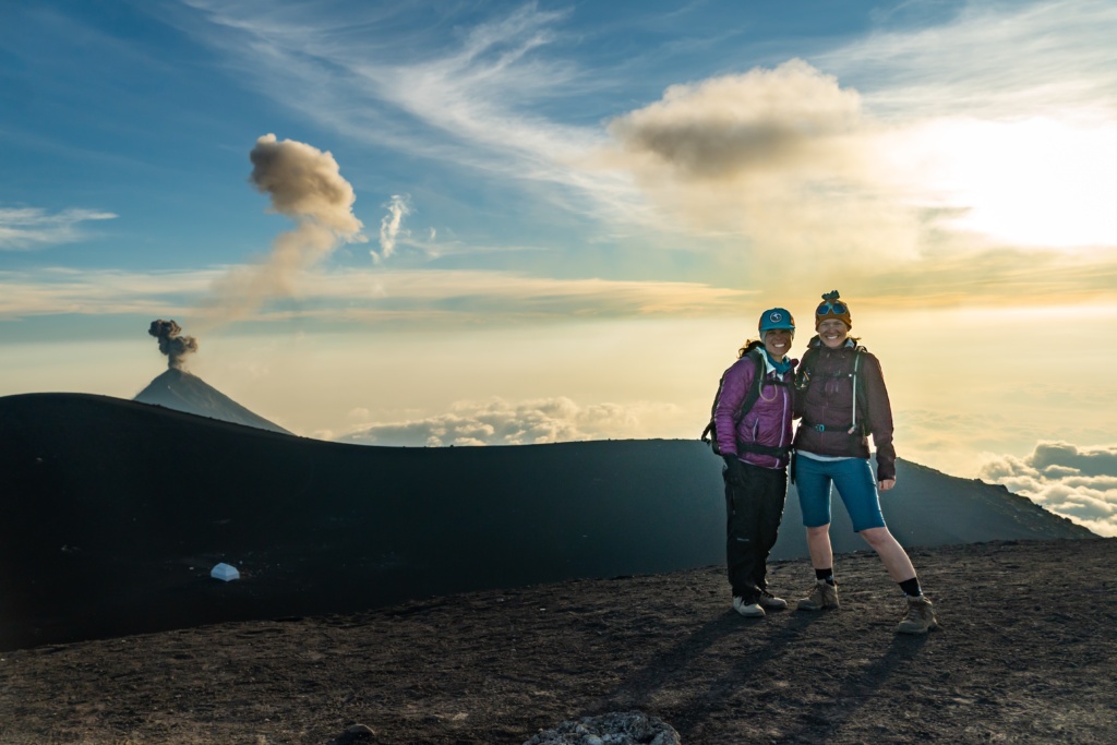 Two mountain bikers stand in front of a smoking volcano in Guatemala