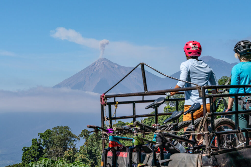Two mountain bikers in the back of a truck looking at a volcano in the distance