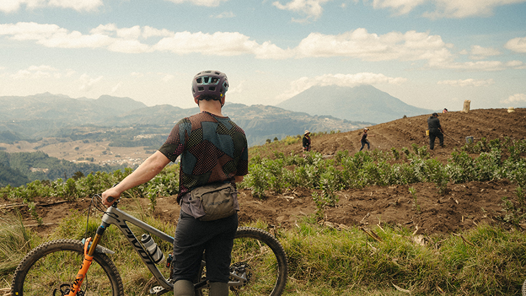 Brice stands next to a bike with his back to the camera. He is looking out at a field in Guatemela